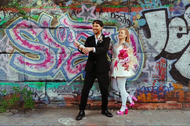 retro styled wedding couple in front of graffiti wall popping champagne bottle