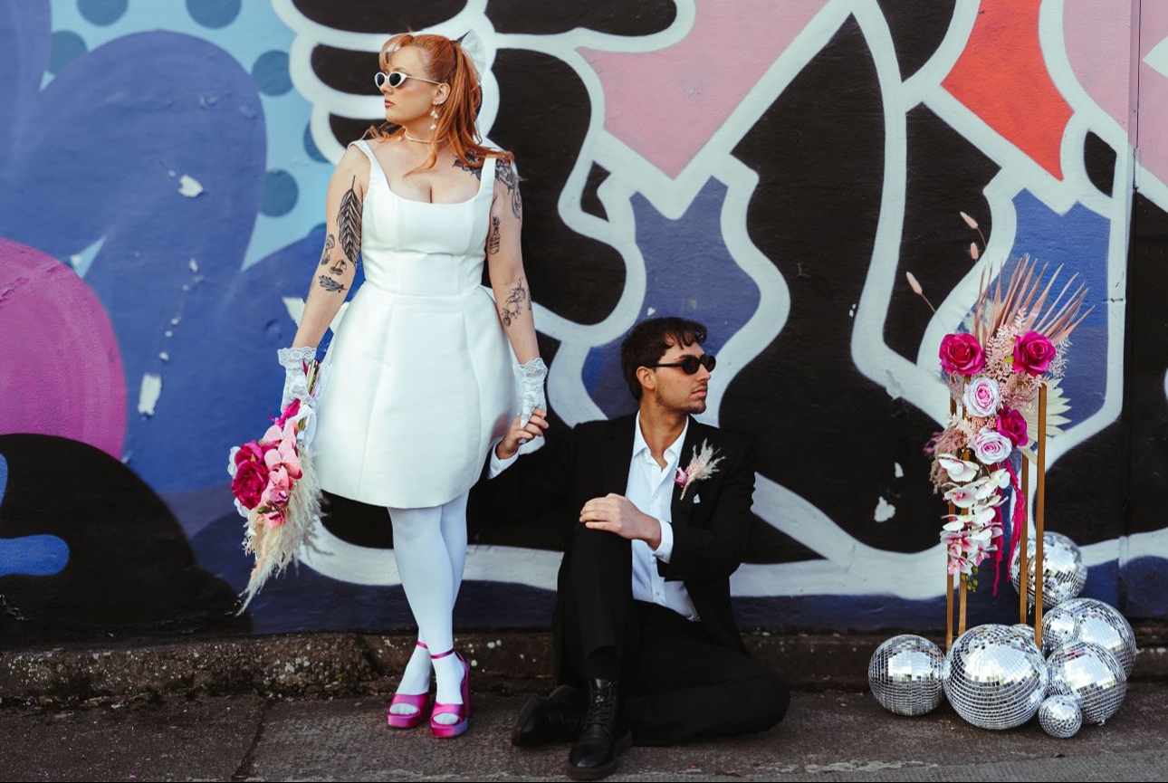 retro styled wedding couple in front of graffiti wall groom sat down bride standing up